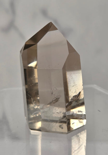 A natural smoky quartz crystal point with internal inclusions, displayed on a clear acrylic stand against a light gray marble background.
