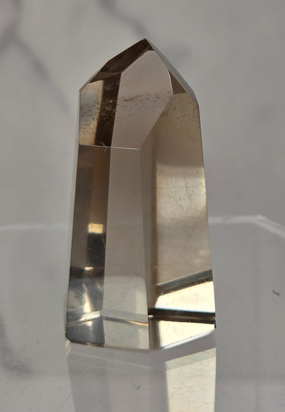 A natural smoky quartz crystal point tower displayed on a clear acrylic stand against a light gray marble-patterned background.