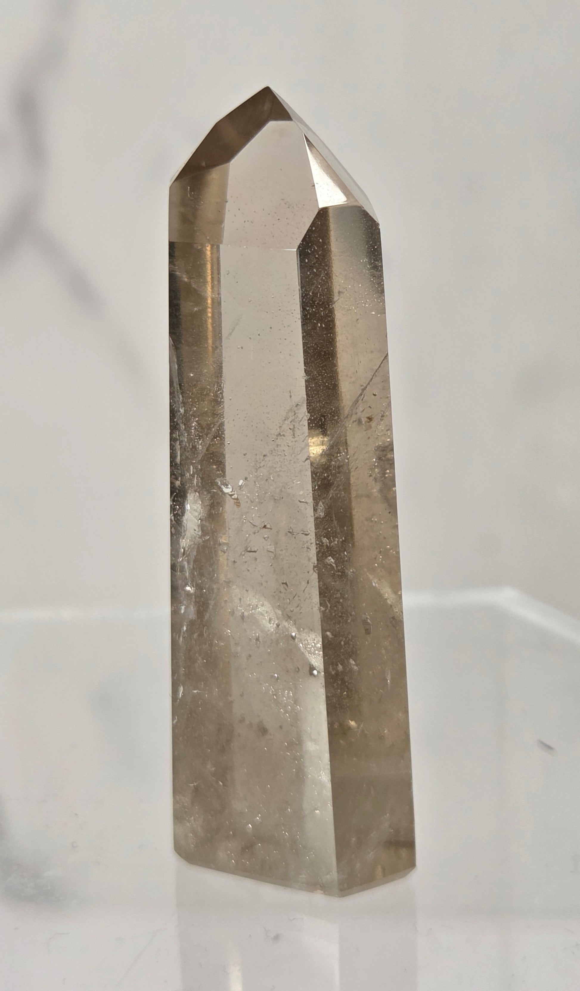 A natural smoky quartz crystal point with a hexagonal shape, standing upright on a clear acrylic display stand against a light gray marble-patterned background.