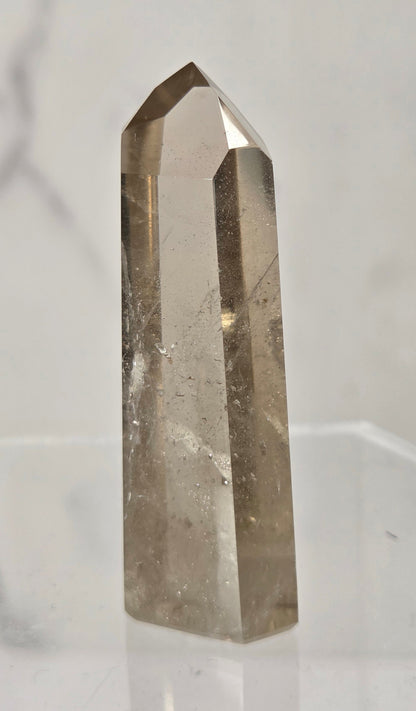 A natural smoky quartz crystal point with a hexagonal shape, standing upright on a clear acrylic display stand against a light gray marble-patterned background.