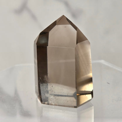 A natural smoky quartz crystal point with a hexagonal shape, resting on a clear acrylic display stand against a light gray marble background.