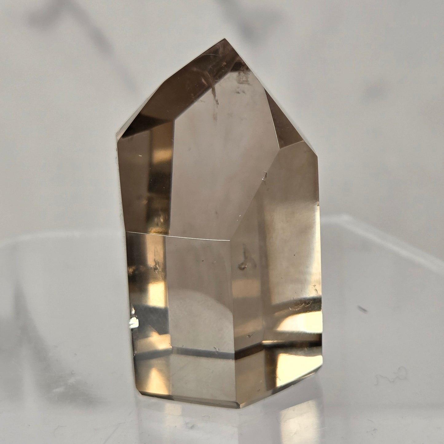 A natural smoky quartz crystal point displayed on a clear acrylic stand against a light gray marble-patterned background.