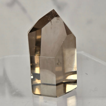 A natural smoky quartz crystal point displayed on a clear acrylic stand against a light gray marble-patterned background.