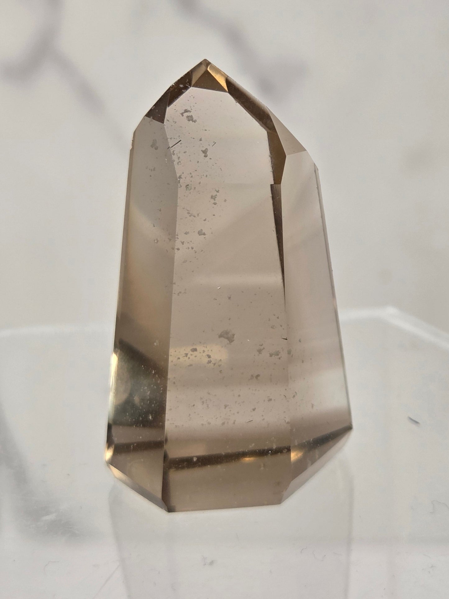 A natural smoky quartz crystal point with a hexagonal shape, standing on a clear acrylic display stand against a light gray marble-patterned background.