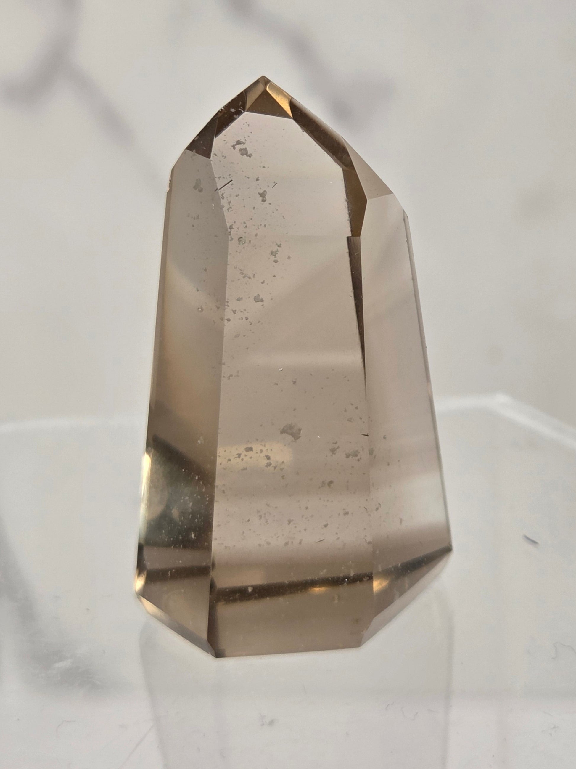 A natural smoky quartz crystal point with a hexagonal shape, standing on a clear acrylic display stand against a light gray marble-patterned background.