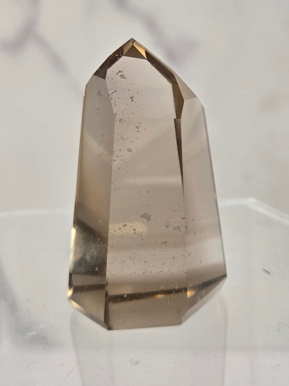 A natural smoky quartz crystal point with a hexagonal shape, standing on a clear acrylic display stand against a light gray marble-patterned background.
