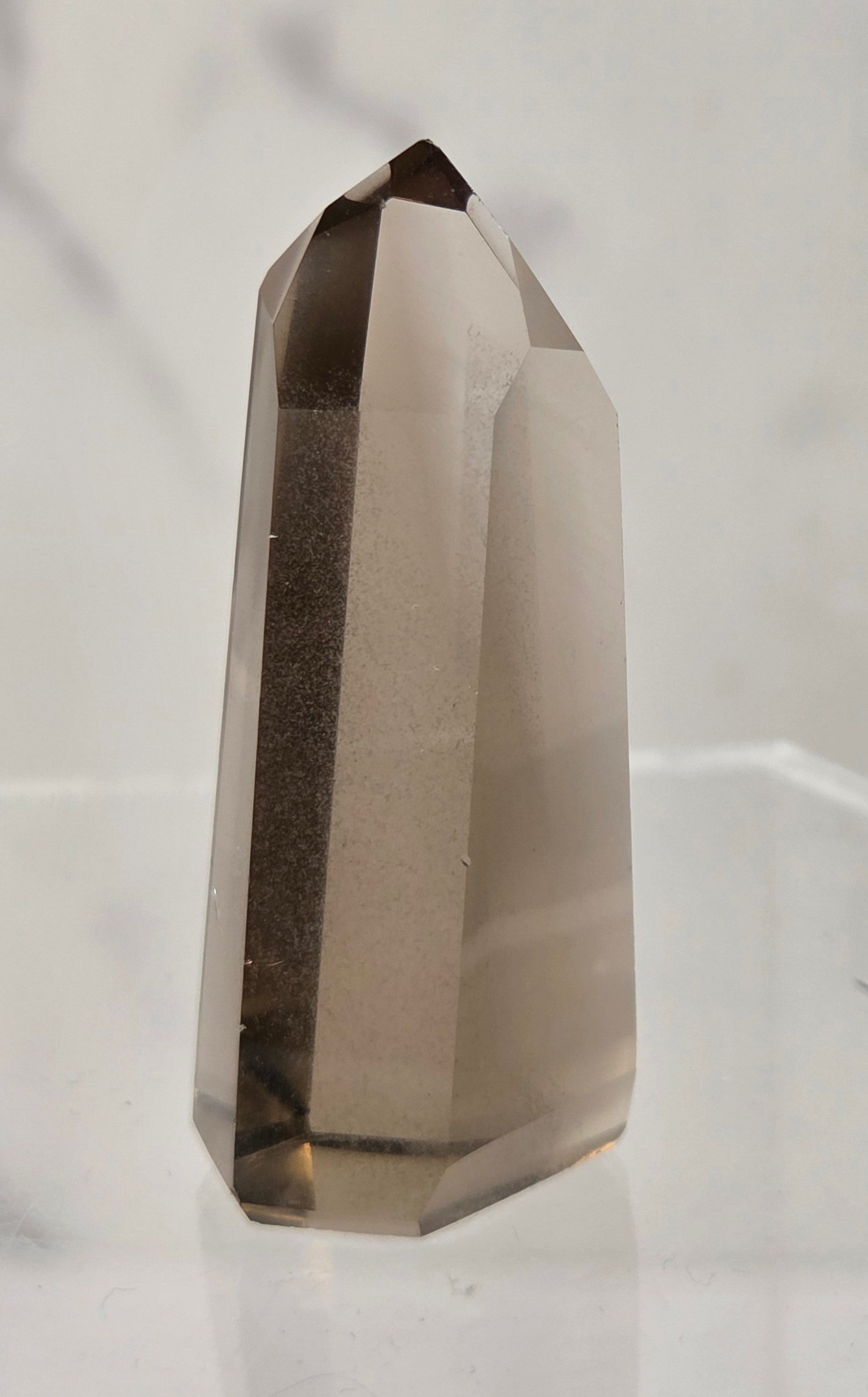 A smoky quartz crystal point with a hexagonal shape, standing upright on a clear acrylic display stand against a light gray marble-patterned background.
