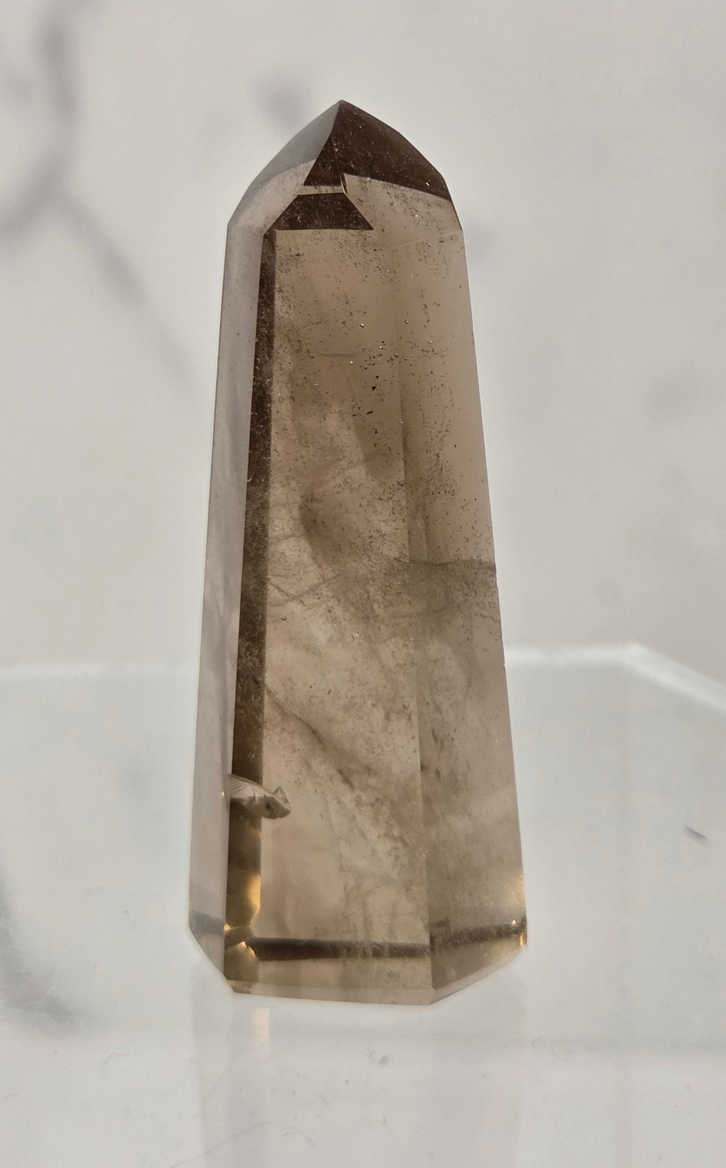 A natural smoky quartz crystal point tower standing upright on a clear acrylic display stand against a light gray marble-patterned background.