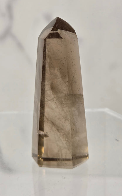 A natural smoky quartz crystal point tower standing upright on a clear acrylic display stand against a light gray marble-patterned background.
