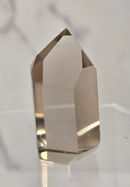 A faceted clear crystal point, likely quartz, standing upright on a transparent acrylic display stand against a light gray marble-patterned background.