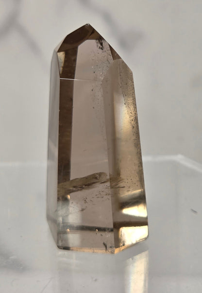 A natural smoky quartz crystal point standing upright on a clear acrylic display block against a light gray marble-patterned background.