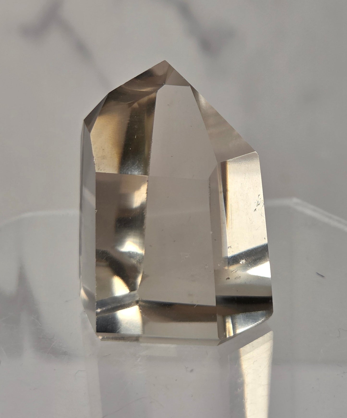 A clear, faceted crystal point mineral specimen standing upright on a transparent acrylic display stand against a light gray marble-patterned background.