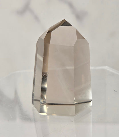 A faceted clear quartz crystal tower with a pointed top, displayed on a transparent acrylic stand against a light gray marble-patterned background.