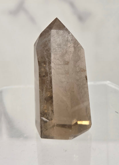 A natural smoky quartz crystal point standing upright on a clear acrylic display stand against a light gray marble-patterned background.