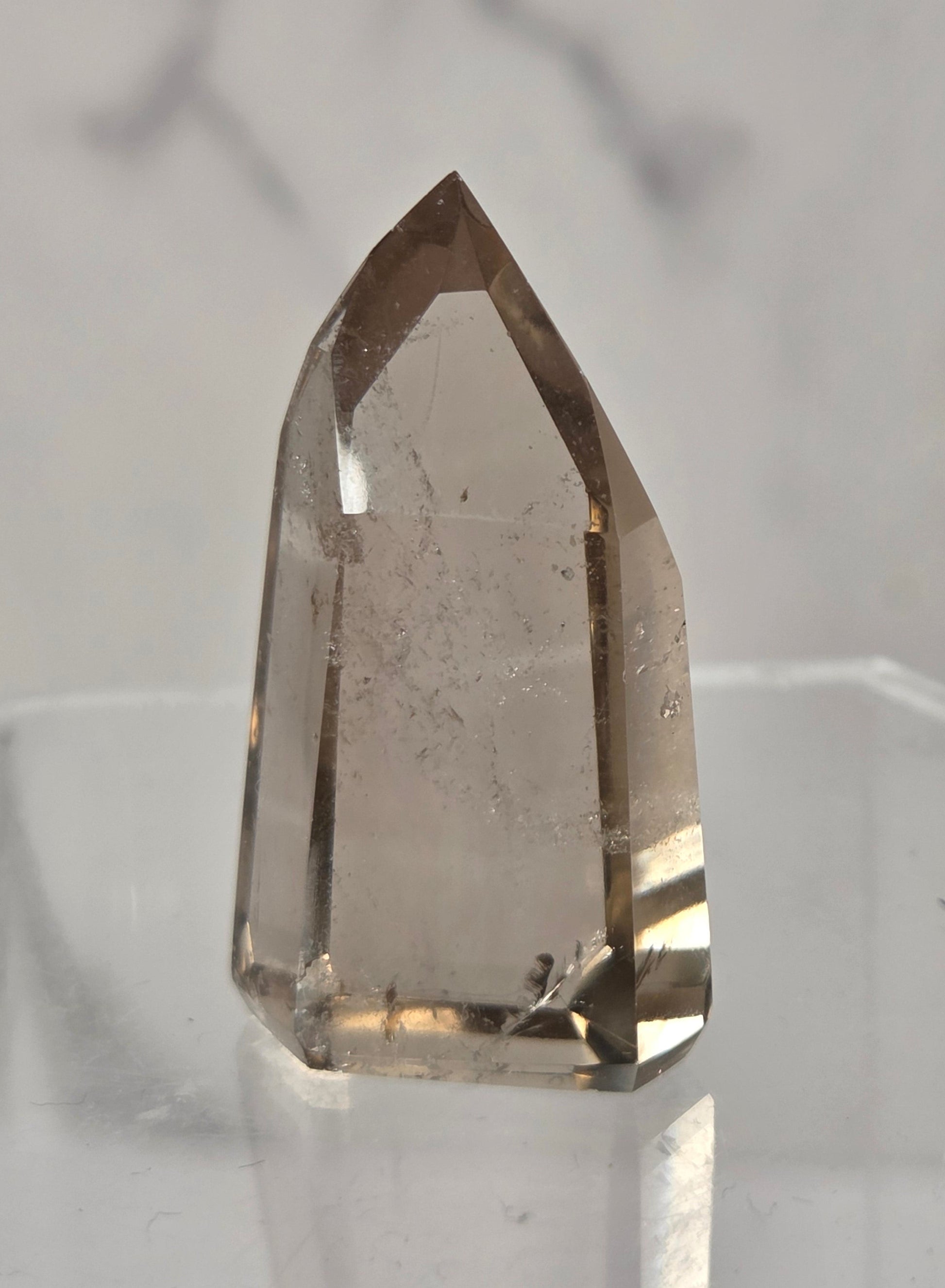 A natural smoky quartz crystal point with internal inclusions, displayed on a clear acrylic stand against a light gray marble background.