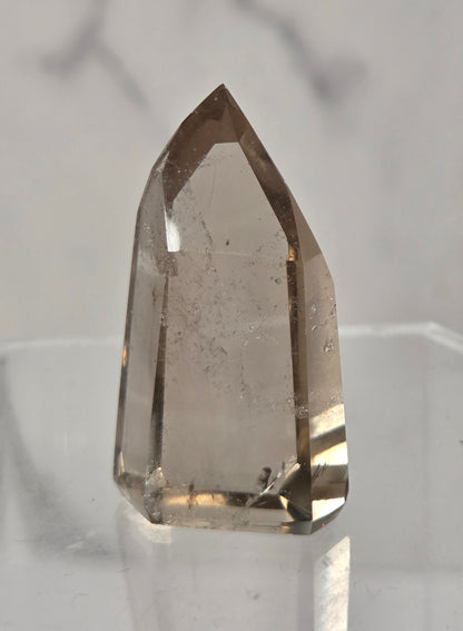 A natural smoky quartz crystal point with internal inclusions, displayed on a clear acrylic stand against a light gray marble background.