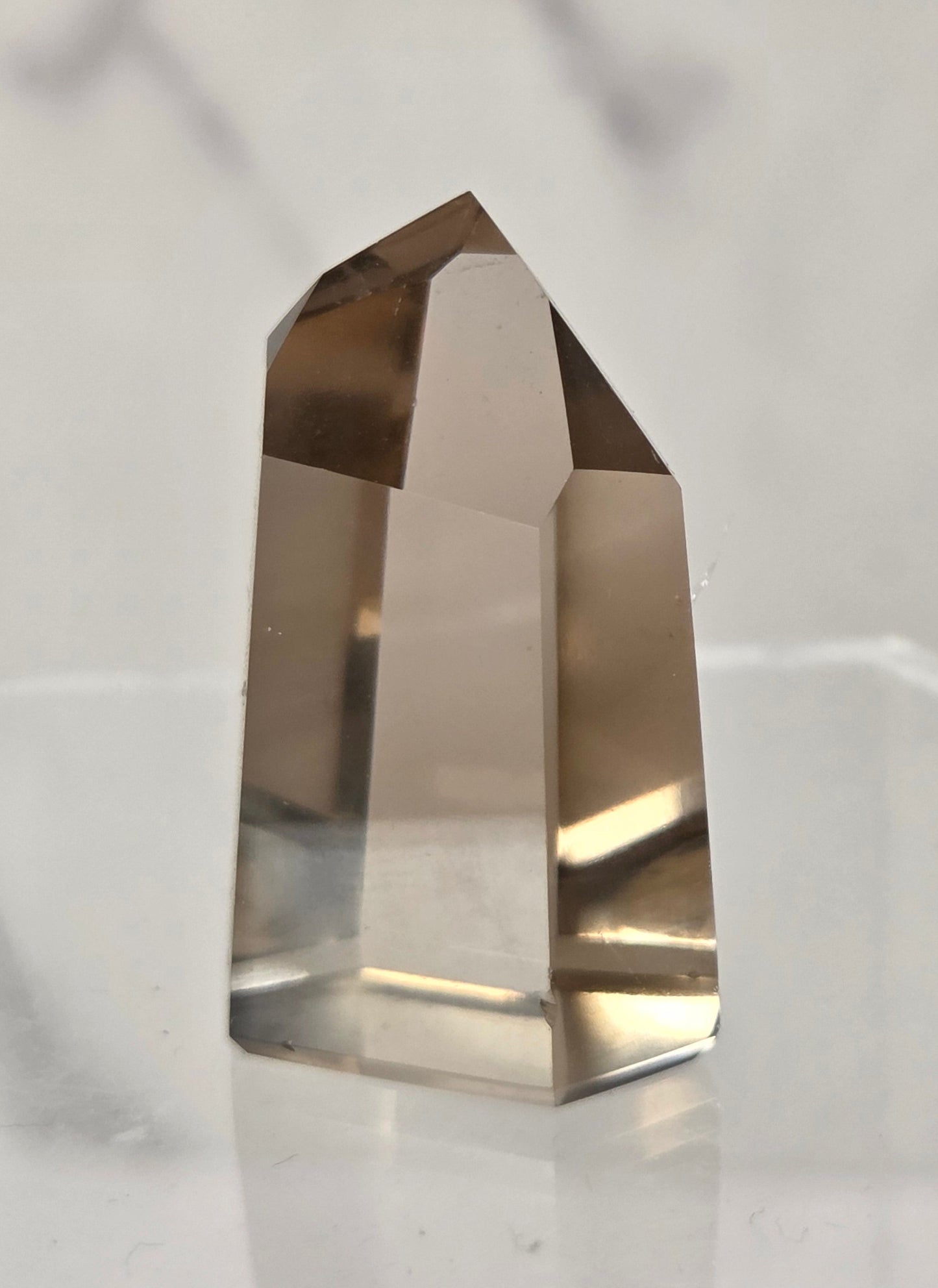 A faceted smoky quartz crystal point displayed on a clear acrylic stand against a light gray marble-patterned background.