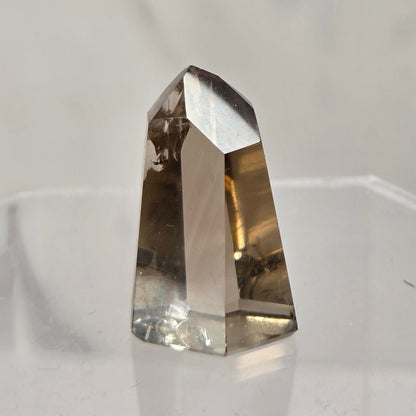A natural smoky quartz crystal point with a translucent brown and smoky gray color, displayed on a clear acrylic stand against a light gray marble-patterned background.