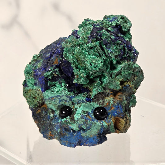 azurite and malachite specimen with a face of black eyes and carved mouth on a clear stand against a white background