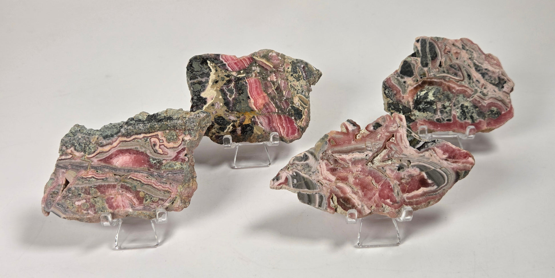 Four raw mineral or stone specimens with pink, gray, black, and green marbling, each displayed on a small clear acrylic stand against a white surface and light gray marble-look background.