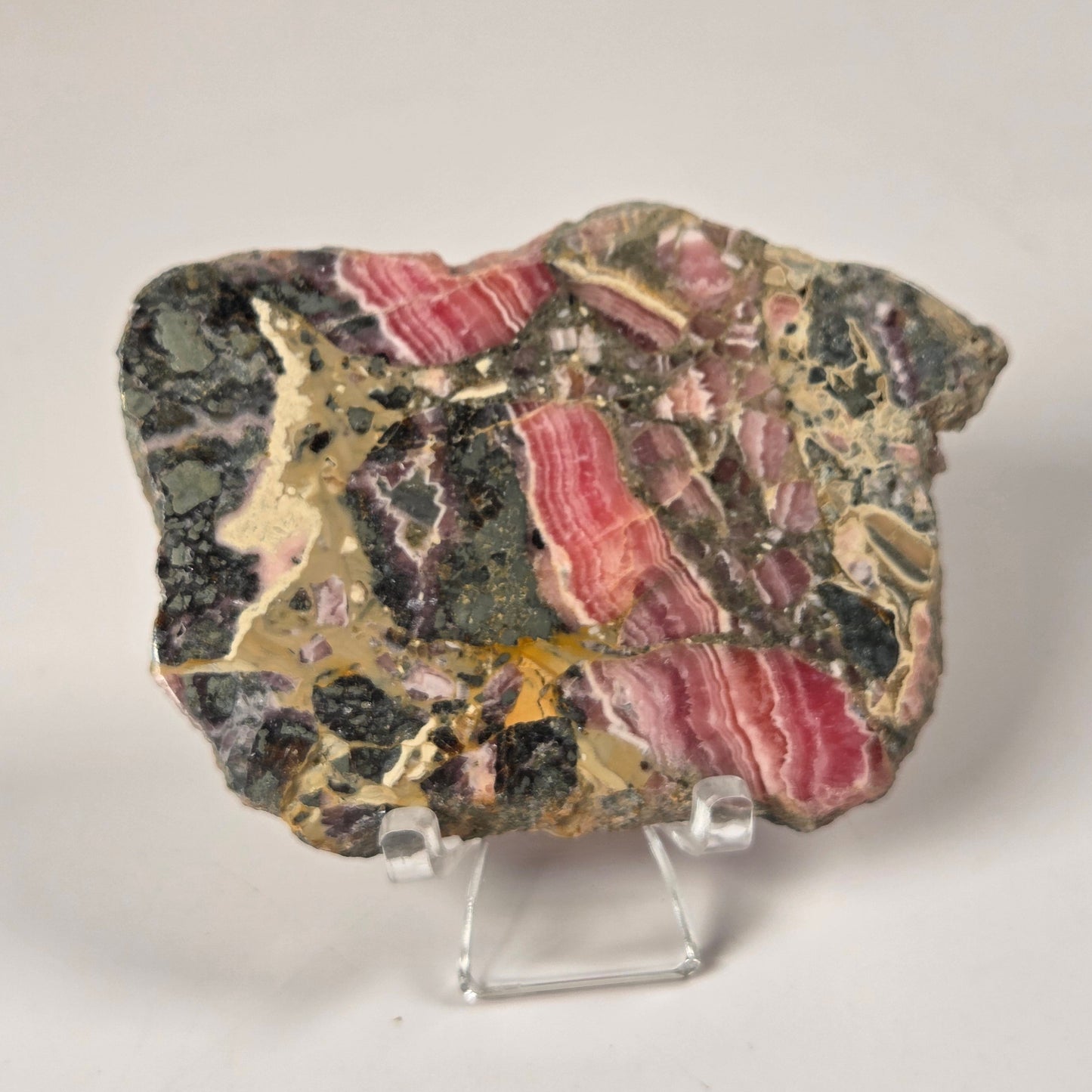 A polished mineral specimen with pink, green, black, and gold banded patterns, displayed on a small clear acrylic stand against a neutral light gray background.