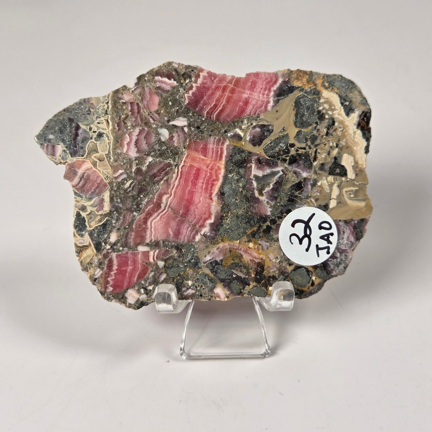 A polished mineral specimen with prominent pink banded bands over a dark gray and black matrix, displayed on a clear plastic stand against a light gray background.