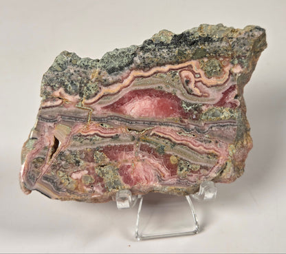 A polished pink and multicolored agate stone slab with layered bands of red, pink, gray, and green, displayed on a clear acrylic stand against a neutral light gray background.