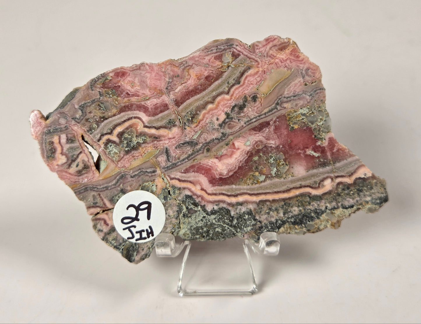 A polished stone slab with layered pink, red, gray, and green bands, displayed on a clear acrylic stand against a light gray background. A small white circular sticker with handwritten numbers is attached near the lower left edge.