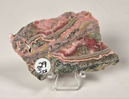 A polished stone slab with layered pink, red, gray, and green bands, displayed on a clear acrylic stand against a light gray background. A small white circular sticker with handwritten numbers is attached near the lower left edge.