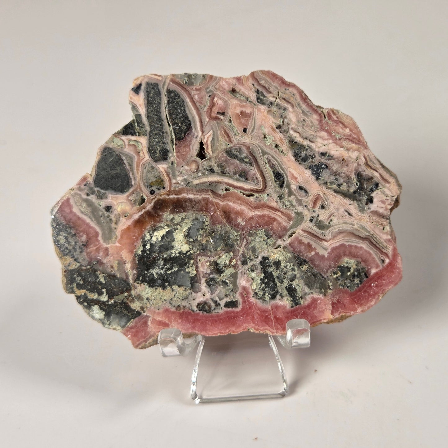 A polished mineral or stone slab with layered pink, black, gray, and pale green bands, displayed on a small clear acrylic stand against a plain light background.