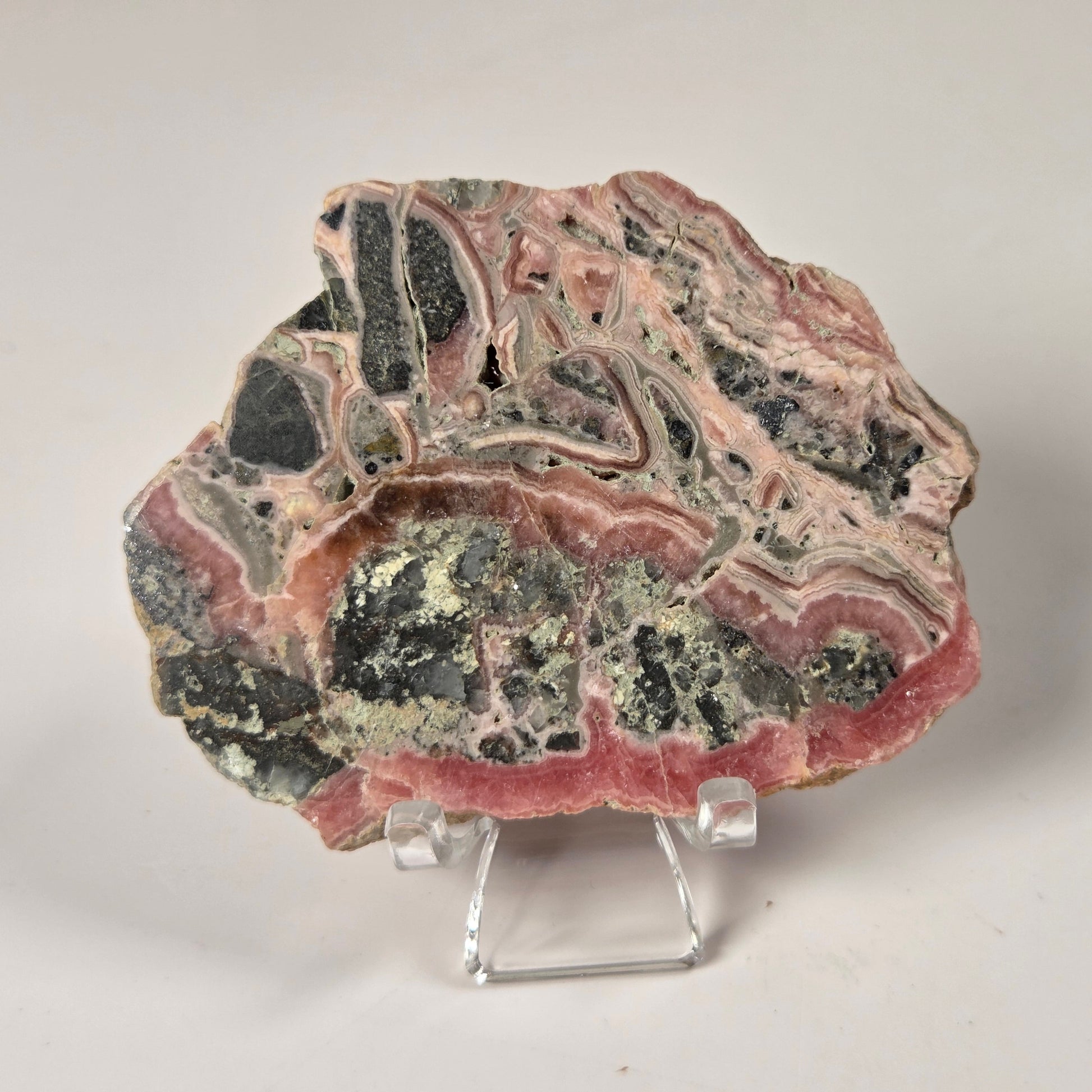 A polished mineral or stone slab with layered pink, black, gray, and pale green bands, displayed on a small clear acrylic stand against a plain light background.
