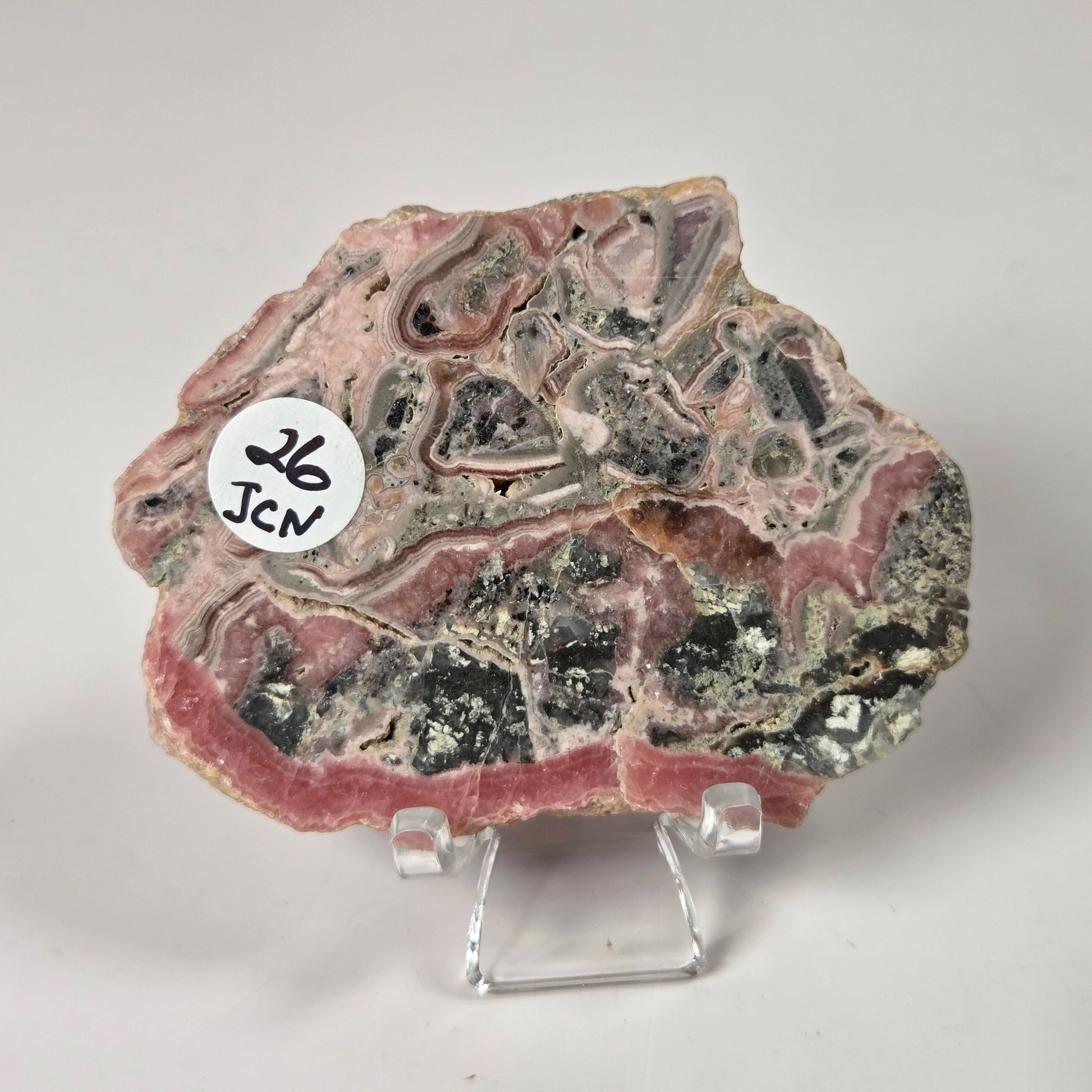 A polished mineral or geode slab with layered pink, gray, black, and metallic patterns, displayed on a small clear acrylic stand against a light gray background. A white circular sticker with the text 'JCN 26' is attached to the left side.