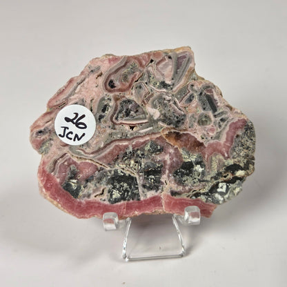 A polished mineral or geode slab with layered pink, gray, black, and metallic patterns, displayed on a small clear acrylic stand against a light gray background. A white circular sticker with the text 'JCN 26' is attached to the left side.