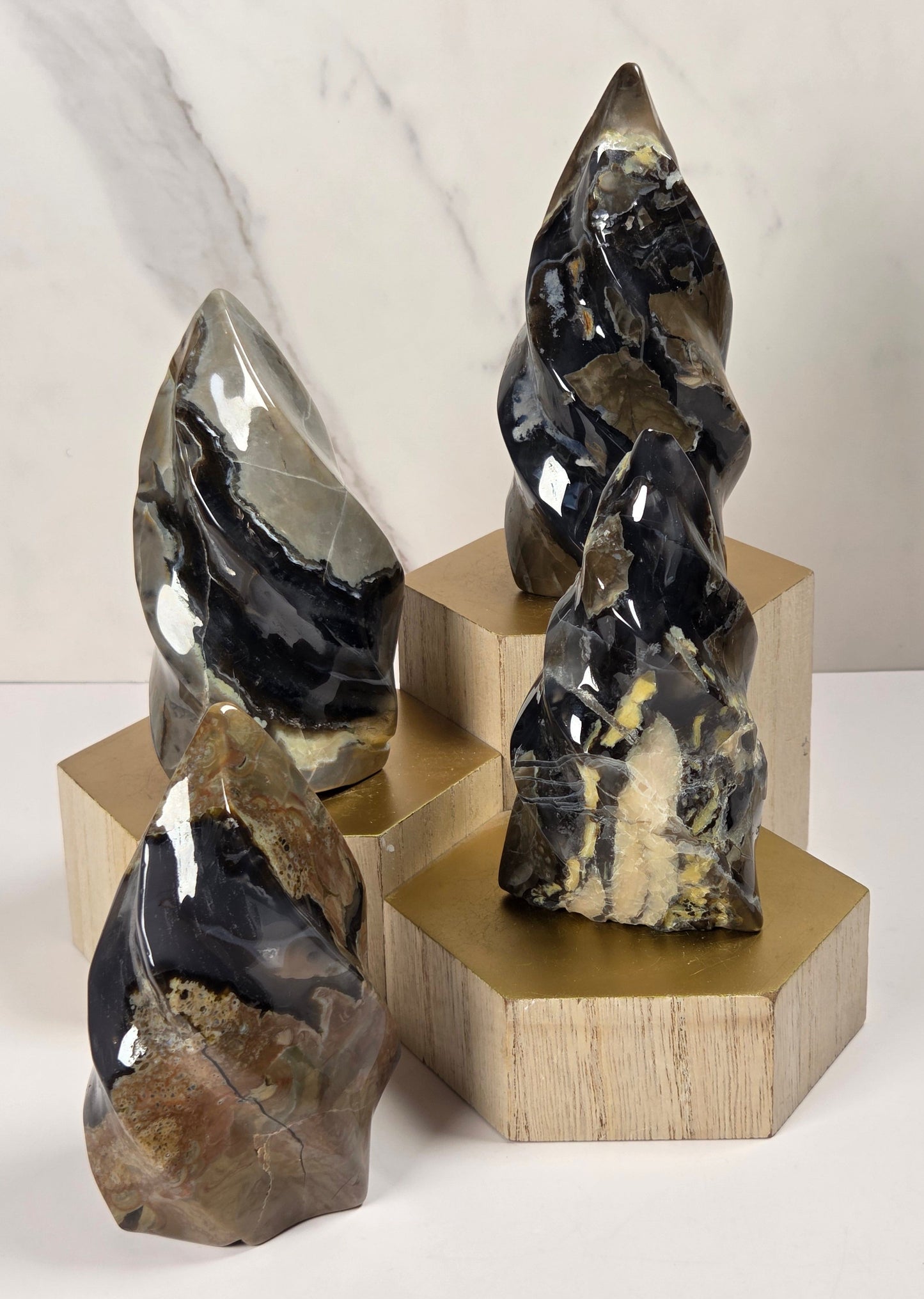 Three polished stone crystal towers in black, gray, and brown with natural banding and textures, displayed on geometric gold and light wood pedestals against a white marble-look background.
