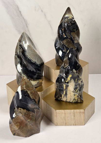 Three polished stone crystal towers in black, gray, and brown with natural banding and textures, displayed on geometric gold and light wood pedestals against a white marble-look background.