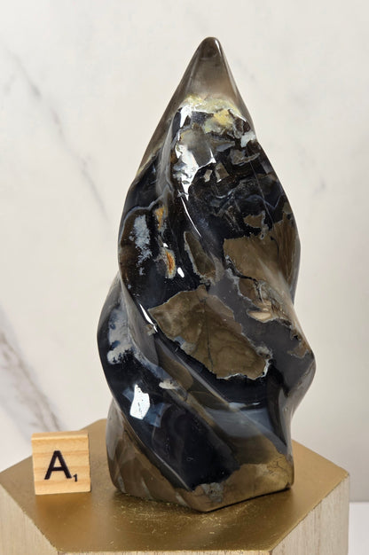 A polished abstract mineral sculpture with swirling black, gray, and metallic gold patterns, displayed on a gold hexagonal pedestal next to a small wooden letter block.