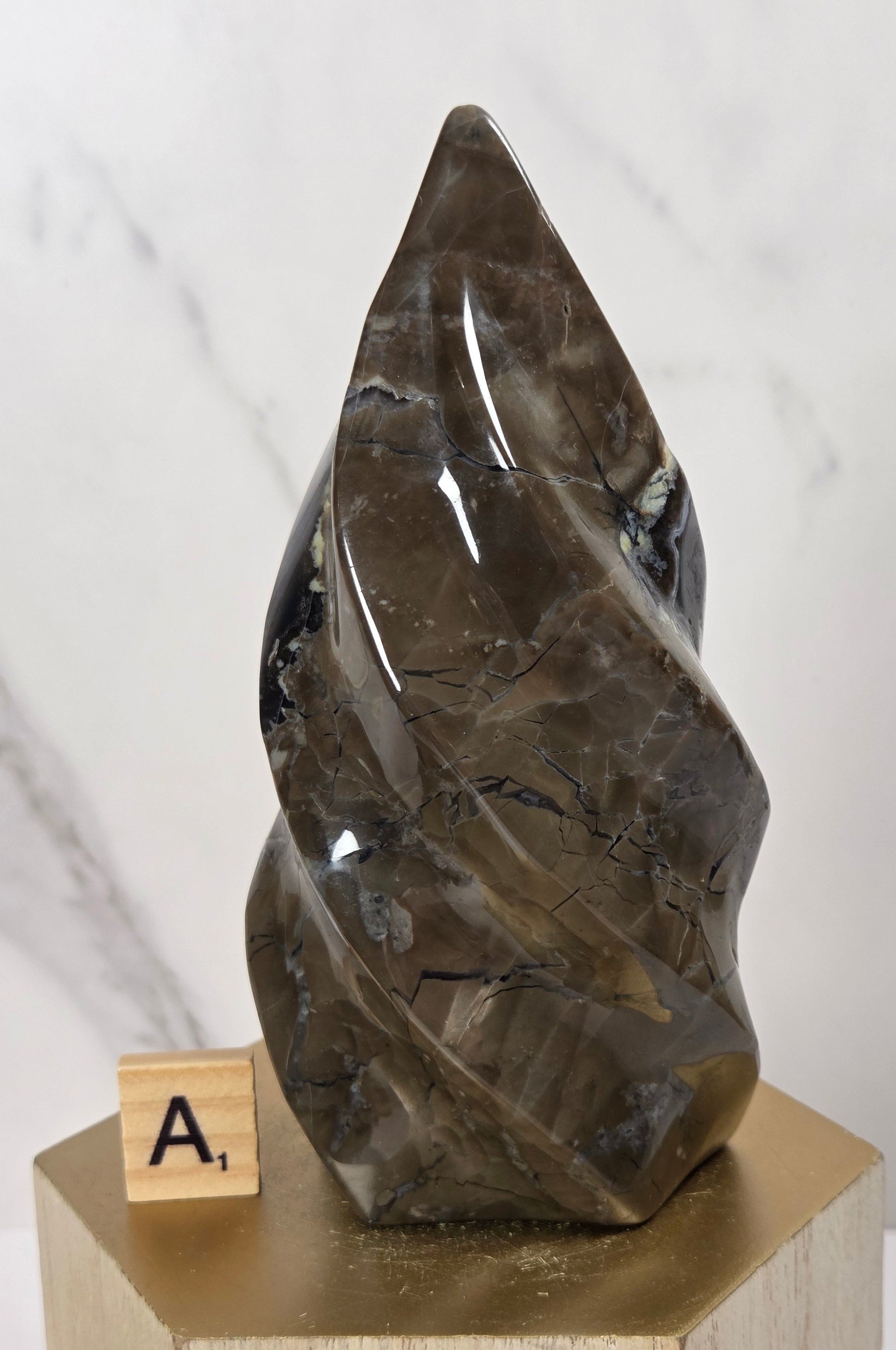 A polished dark brown smoky quartz crystal sculpture with natural inclusions, displayed on a light wood pedestal next to a Scrabble tile on a white marble background.