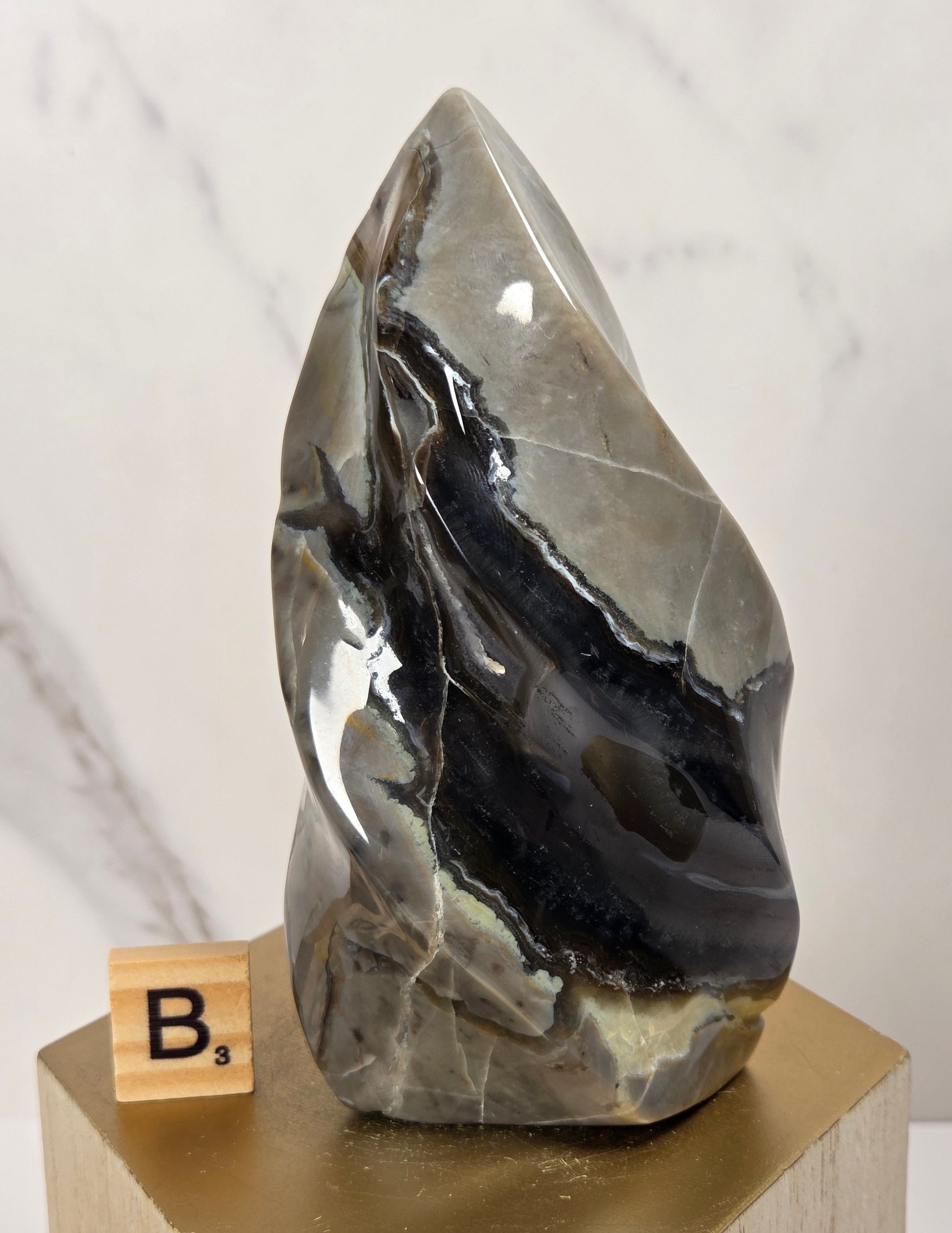 A polished natural stone sculpture with swirling black, gray, and beige bands, displayed on a gold hexagonal pedestal next to a small Scrabble tile.