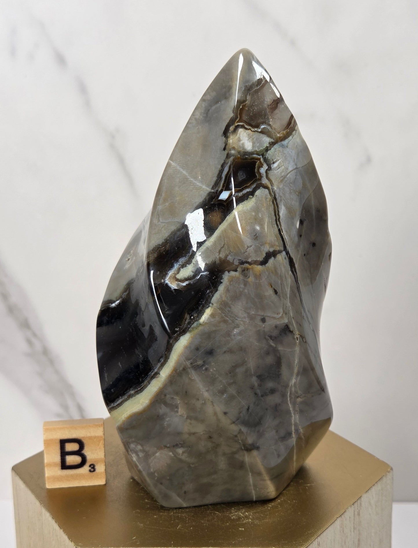 A polished natural stone mineral specimen with dark gray, black, and beige veining, displayed on a gold-colored geometric pedestal with a small wooden letter block showing 'B3' next to it, set against a light marble-patterned background.