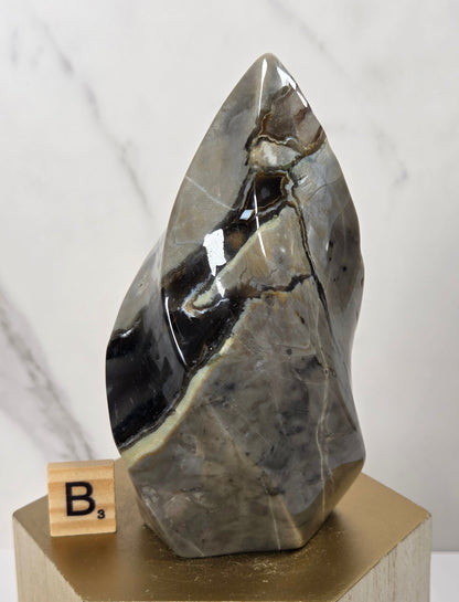 A polished natural stone mineral specimen with dark gray, black, and beige veining, displayed on a gold-colored geometric pedestal with a small wooden letter block showing 'B3' next to it, set against a light marble-patterned background.