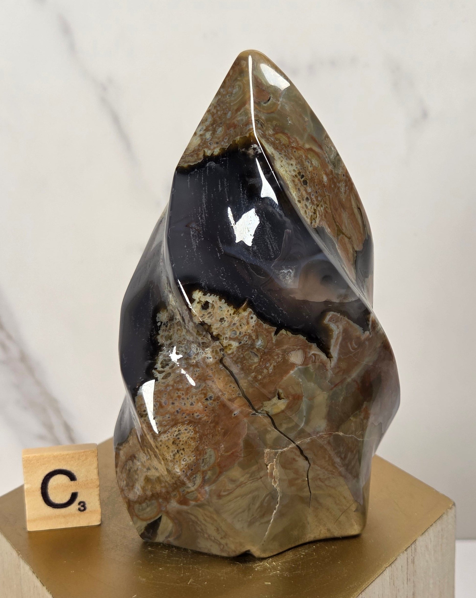 A polished natural crystal or geode specimen with layered brown, tan, and dark black sections, displayed on a gold-colored geometric pedestal next to a small Scrabble tile showing the letter C-5.