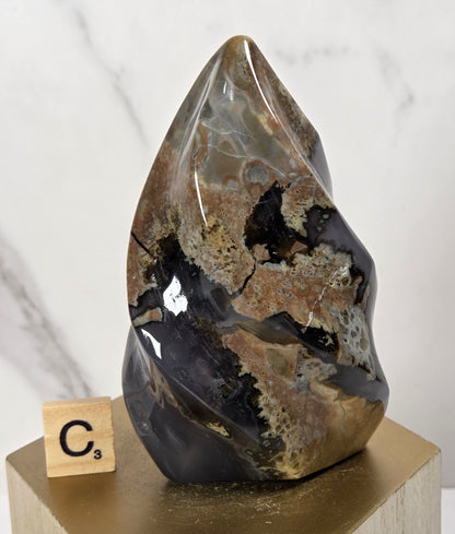 A polished natural crystal or mineral specimen with a teardrop shape, featuring brown, black, and gray marbled patterns, displayed on a light wood block next to a small Scrabble tile on a gold-colored pedestal.