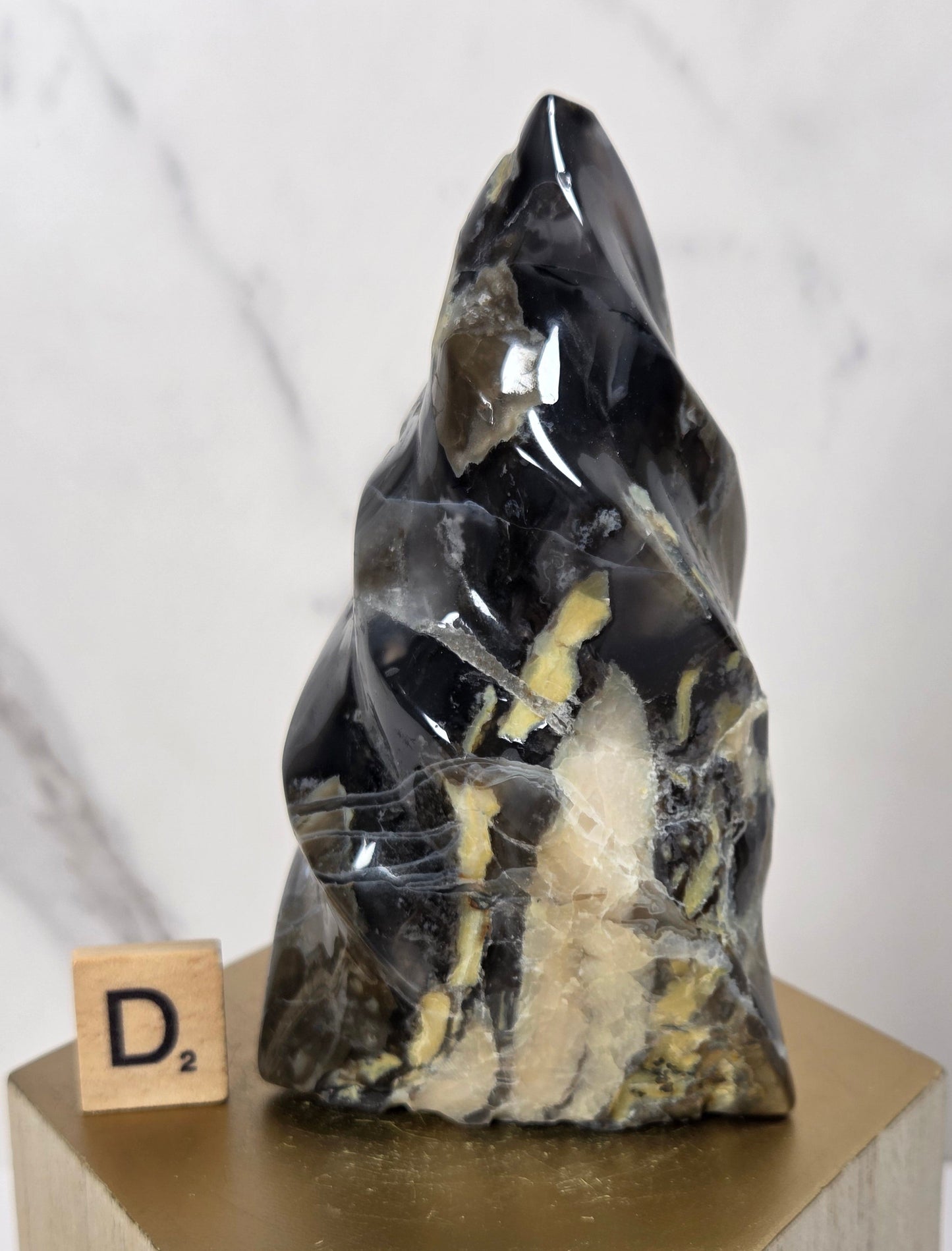 A polished black crystal mineral specimen with prominent gold and beige matrix inclusions, displayed on a gold-colored geometric pedestal next to a wooden Scrabble tile.