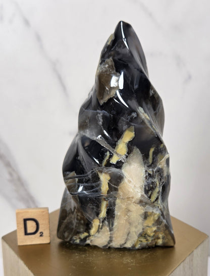 A polished black crystal mineral specimen with prominent gold and beige matrix inclusions, displayed on a gold-colored geometric pedestal next to a wooden Scrabble tile.
