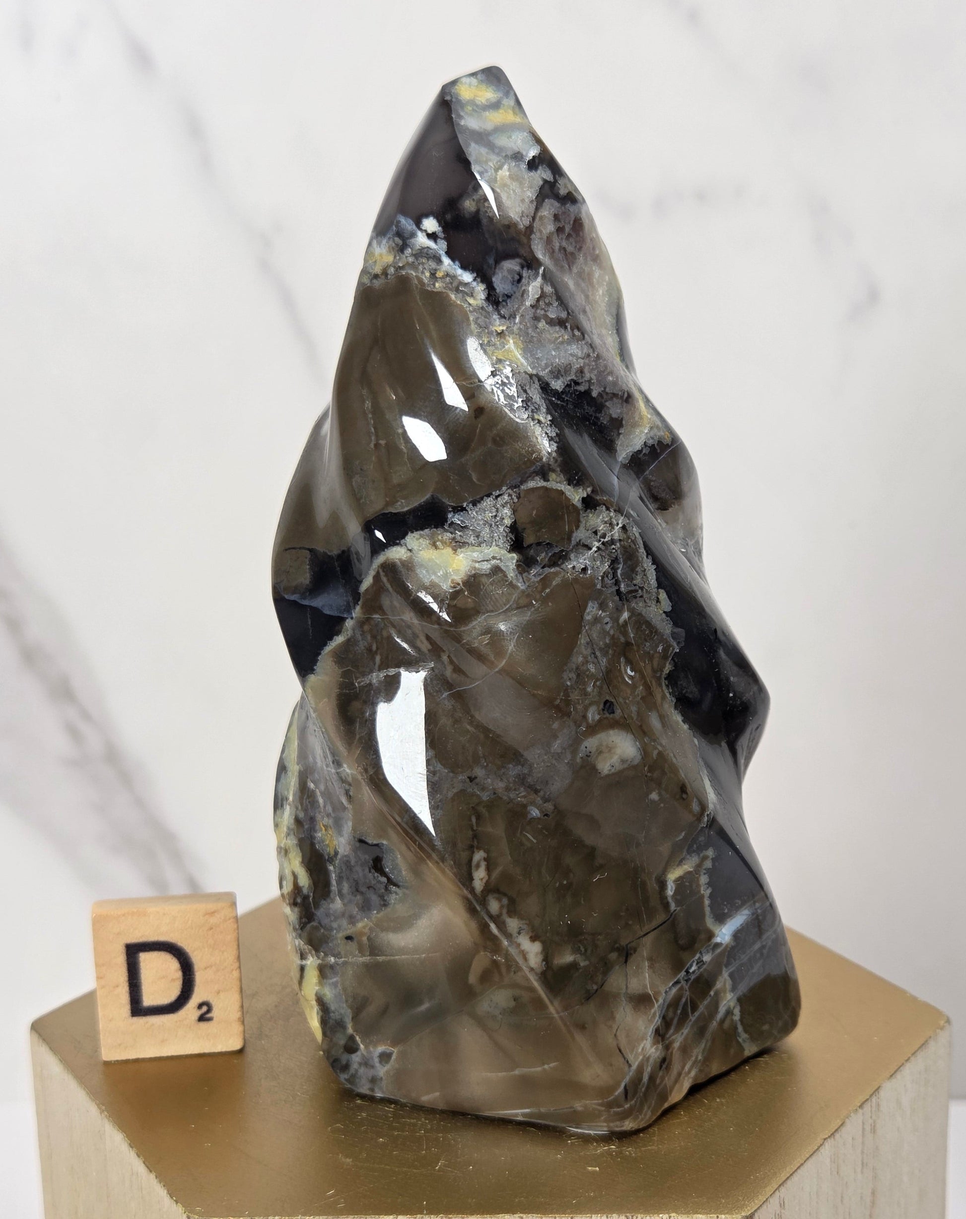 A polished natural crystal or mineral sculpture with a pointed top, displayed on a light wood block pedestal. The crystal is dark gray with black and metallic gold veining, and a small wooden letter tile spelling 'D2' sits beside it.