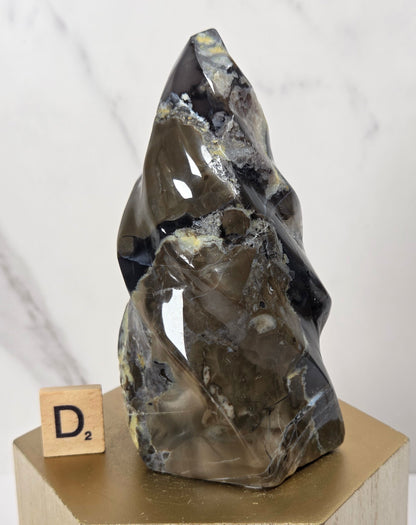 A polished natural crystal or mineral sculpture with a pointed top, displayed on a light wood block pedestal. The crystal is dark gray with black and metallic gold veining, and a small wooden letter tile spelling 'D2' sits beside it.