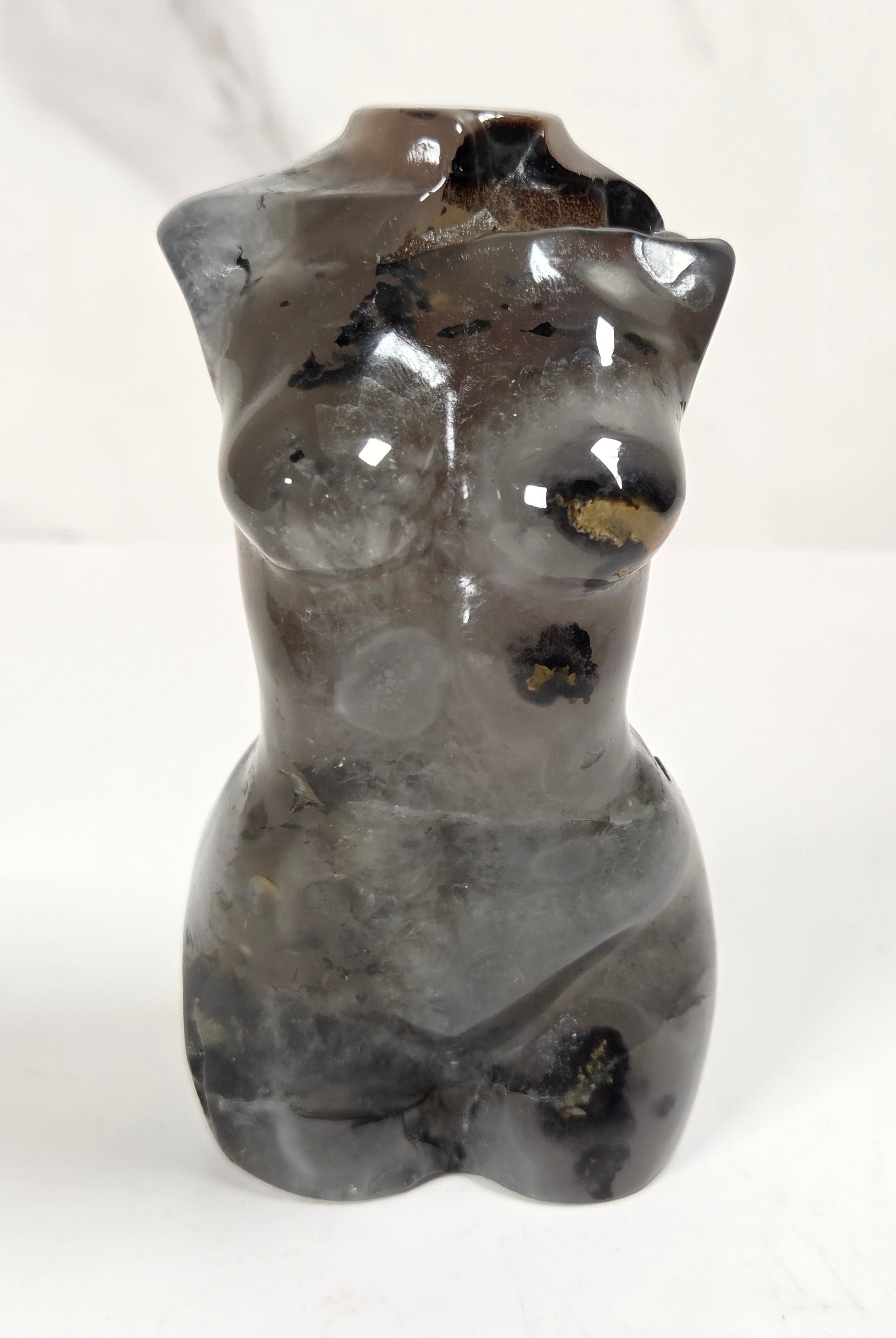 A small sculptural vase shaped like a torso, made from polished dark gray stone with natural black and gold inclusions, set against a light marble-patterned background.