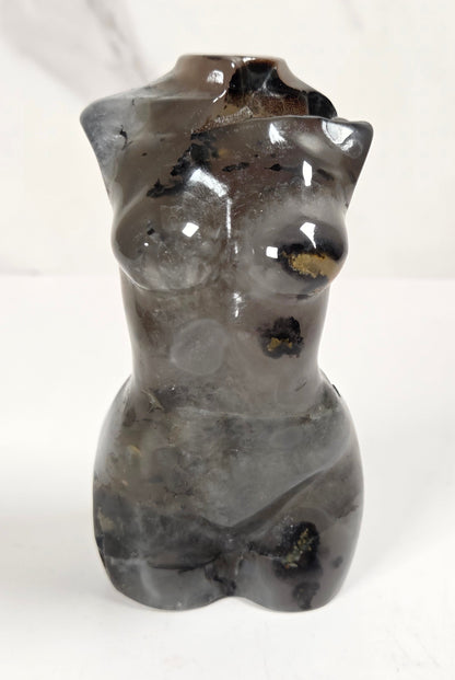 A small sculptural vase shaped like a torso, made from polished dark gray stone with natural black and gold inclusions, set against a light marble-patterned background.