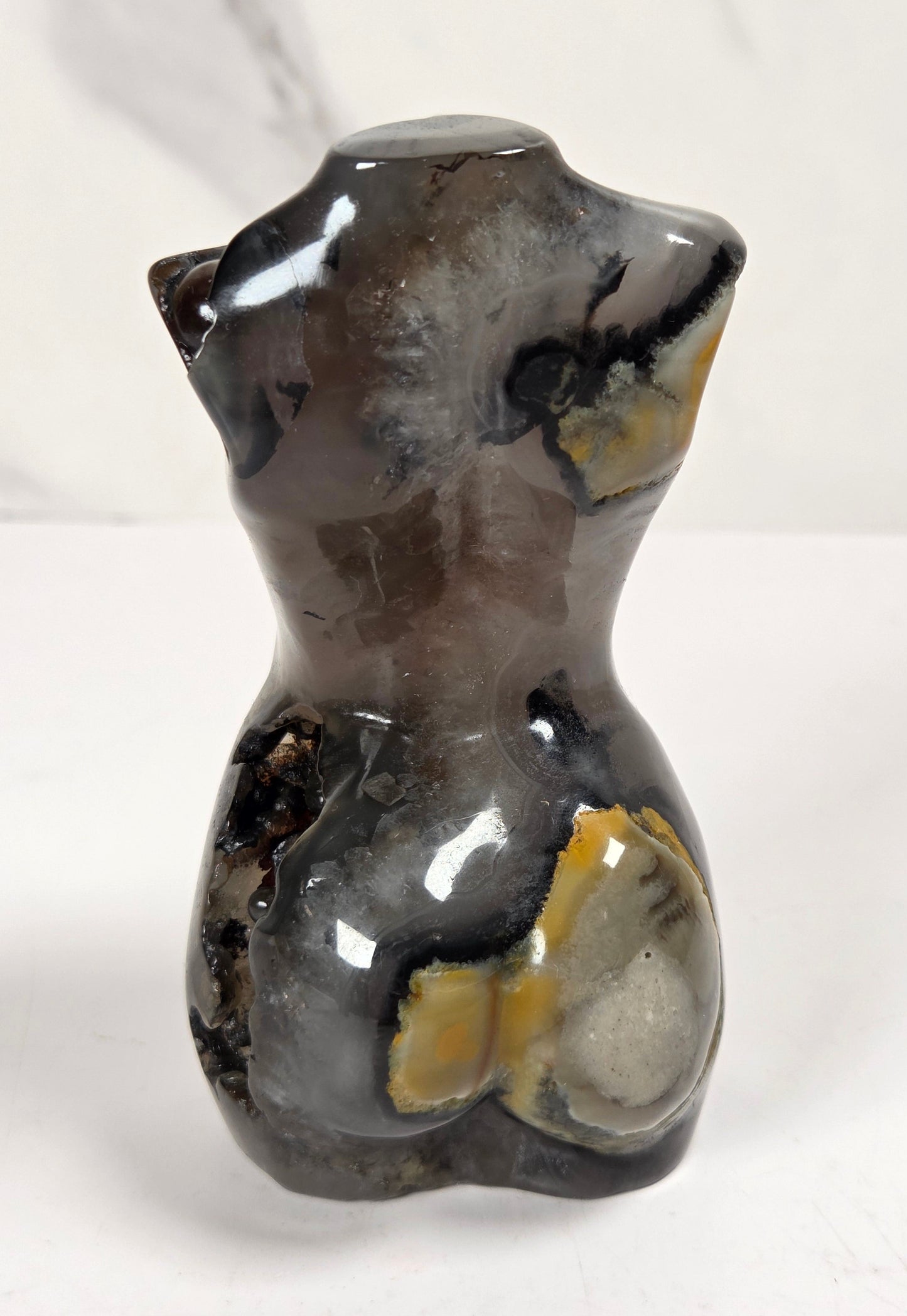 A polished natural stone torso sculpture, likely agate or smoky quartz, featuring dark gray, black, and yellow-brown banding, photographed on a white marble-patterned surface.