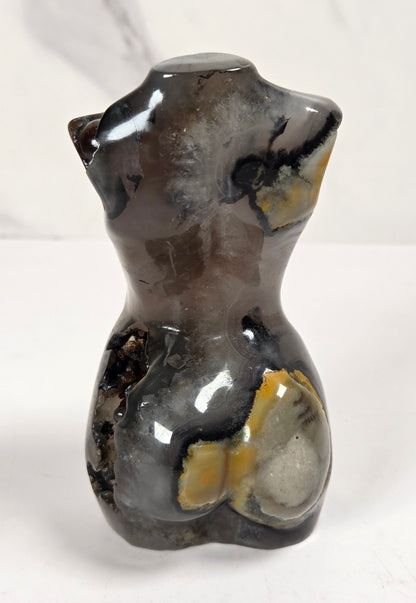 A polished natural stone torso sculpture, likely agate or smoky quartz, featuring dark gray, black, and yellow-brown banding, photographed on a white marble-patterned surface.